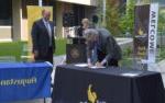 Tim Wynes signing document on table outdoors with 2 people watching and smiling