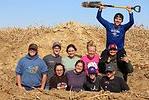 10 students in soil pit with one holding shovel above head
